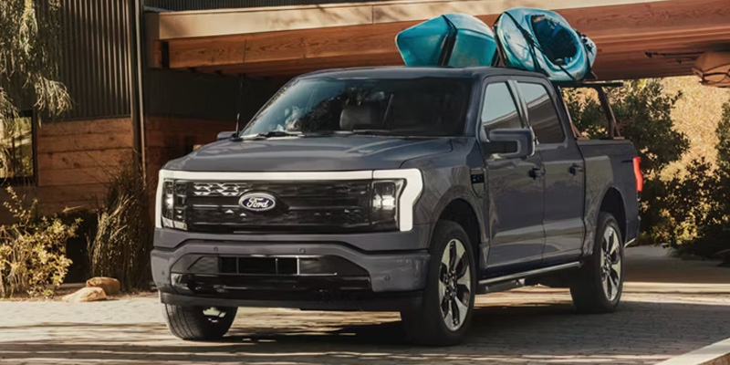 A grey Ford F-150 Lightning electric pickup truck with two blue kayaks on a roof rack, parked in front of a modern wood-accented home.