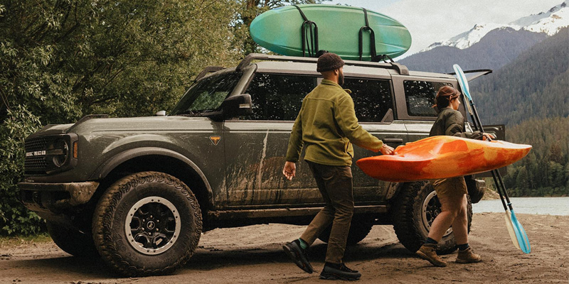 Two people unloading colorful kayaks from a muddy, dark green Ford Bronco parked on a sandy shore, with a scenic lake and pine-covered mountains in the background.