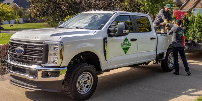 Two workers unloading landscaping supplies from the bed of a white Ford Super Duty truck parked on a residential driveway.
