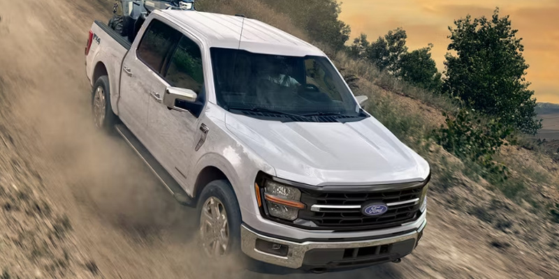 High-angle view of a white Ford F-150 truck kicking up dust as it drives down a steep, rugged dirt incline.