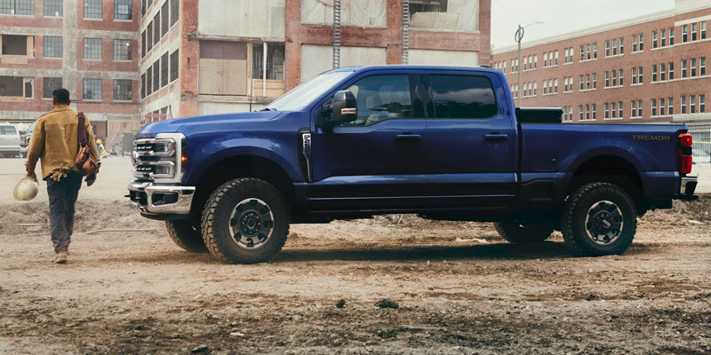 Side profile view of a blue Ford Super Duty F-250 Tremor pickup truck parked at a construction site with a worker holding a hardhat walking past it.