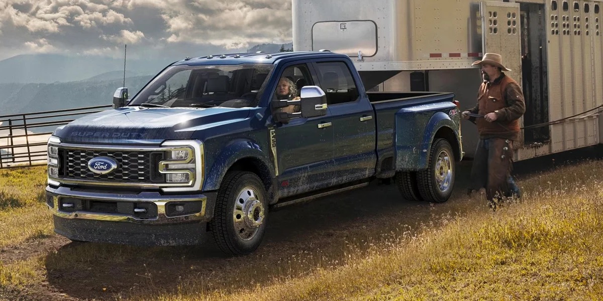 A blue Ford Super Duty dually pickup truck hitched to a large livestock trailer in a rural field.