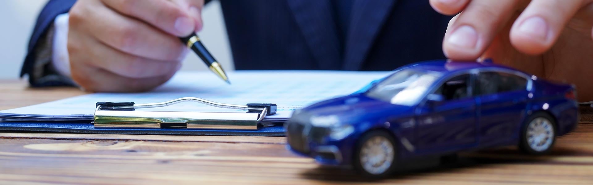 A close-up of a person holding a pen over contract paperwork on a clipboard while resting a hand on a miniature blue toy sedan on a wooden desk.