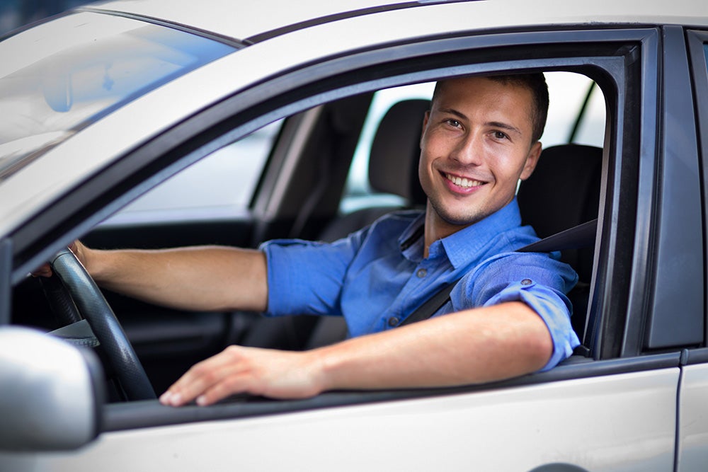 A smiling man in a blue button-down shirt resting his arm on the open window of a silver car.
